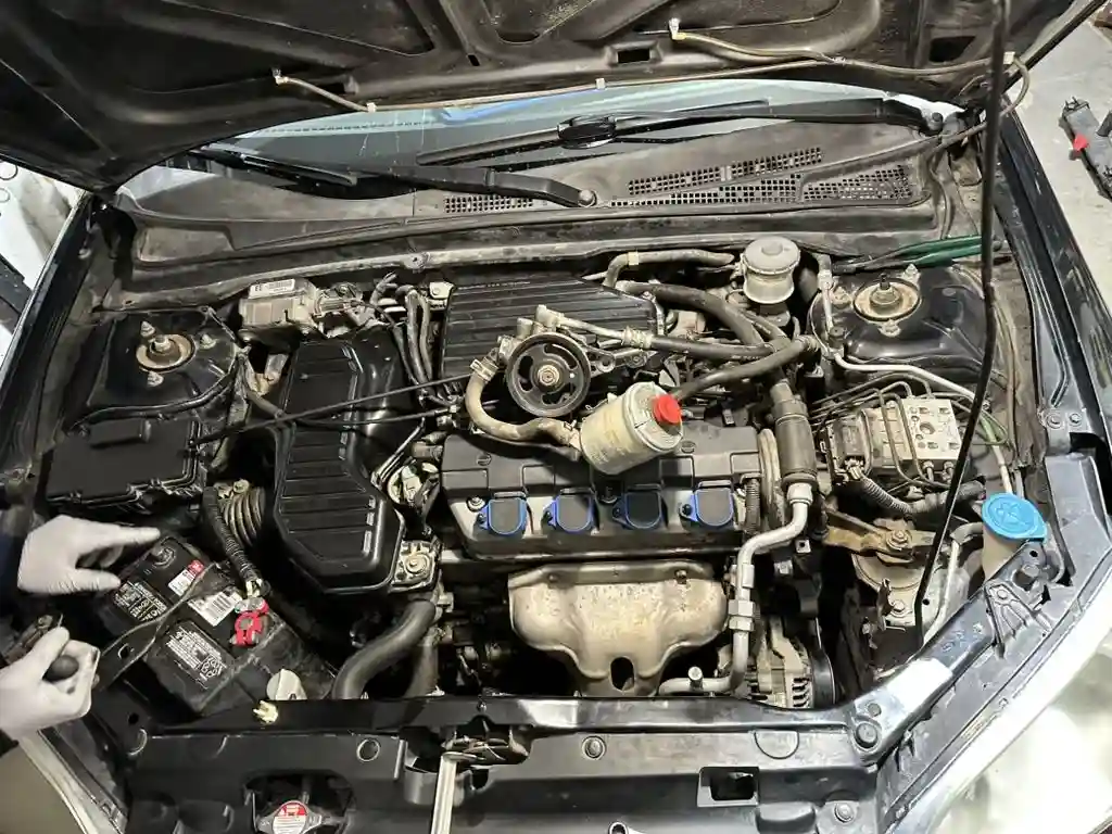A Honda Civic engine bay with the hood up, as a mechanic in gloves points at the battery